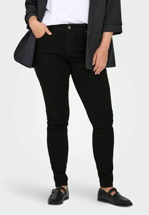 Jean skinny en denim noir avec une silhouette ajustée, dotés d'une fermeture à bouton à l'avant et de poches arrière. Associé à un haut blanc et à un blazer sombre.
