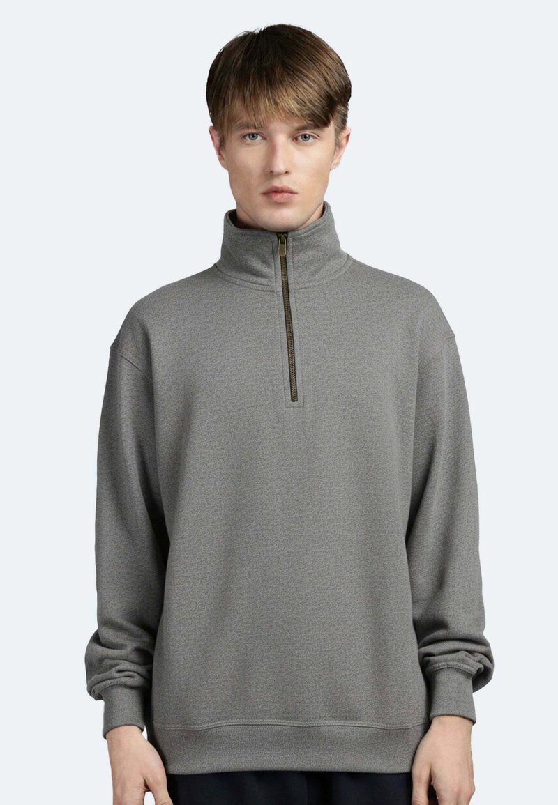 Junger Mann trägt einen grauen Pullover mit Viertel-Reißverschluss und langen Ärmeln, steht vor einem schlichten hellen Hintergrund.