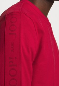 Primer plano de una persona que lleva un sudadera roja brillante con el texto "Joop! Jeans" bordado en la franja del hombro.
