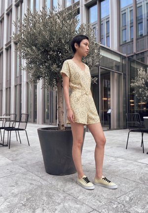Jeune femme debout près d’un arbre en pot dans un espace urbain en plein air, portant une combinaison beige fleurie et des baskets claires avec des lacets noirs.