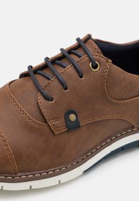 Chaussure en cuir brun avec des lacets noirs, des accents perforés et une semelle blanche en contraste. Présente un rivet métallique décoratif et des détails de couture.