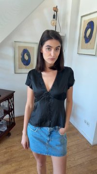 Γυναίκα με σκουρόχρωμα μαλλιά που φοράει μαύρο τοπ με κουμπιά και τζιν φούστα στέκεται σε ένα δωμάτιο με ξύλινο δάπεδο και μπλε αφηρημένη ζωγραφική στον τοίχο.