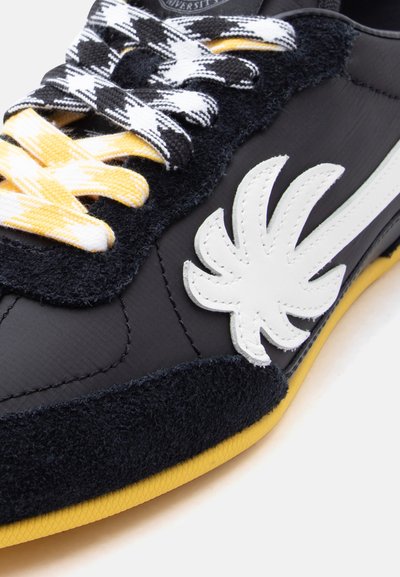 Μαύρο αθλητικό παπούτσι με κίτρινη σόλα, κορδόνια με μοτίβο κίτρινου και ασπρόμαυρου, και λευκό λογότυπο από δερμάτινο σχέδιο σε σχήμα φύλλου στο πλάι.