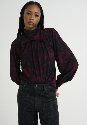 Blusa a maniche lunghe in un motivo astratto nero e bordeaux, con colletto alto e frontale drappeggiato, abbinata a jeans neri decorati con borchie.