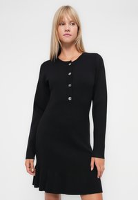 Μαύρο φόρεμα με μακριά μανίκια από υφασμάτινη πλέξη με υφή. Διαθέτει στρογγυλή λαιμόκοψη και πέντε διακοσμητικά κουμπιά κατά μήκος του μπροστινού μέρους. Λεπτομέρεια φαρδιάς στρίφωσης.