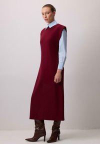 Burgundia ilma varrukatega kleit, mis on valmistatud kudumiskangast, koos helesinise kraega särgiga. Mudel kannab pruunisid põlvektaasiga teravatippe saapaid.