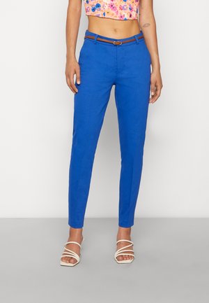 Pantalons ajustés bleu avec une texture lisse, dotés d'une ceinture marron. Les pantalons se rétrécissent au niveau de la cheville, associés à des sandales blanches à lanières.