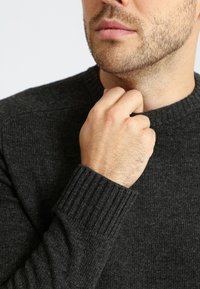 Dunkelgrauer Strickpullover mit gerippten Bündchen und Halsausschnitt, getragen von einer Person, die den Kragen berührt und einen strukturierten Stoff zeigt.