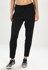 Žena v černých úzkých joggingových kalhotách s elastickým pasem a šedých atletických slip-on teniskách, stojící na bílé podlaze.