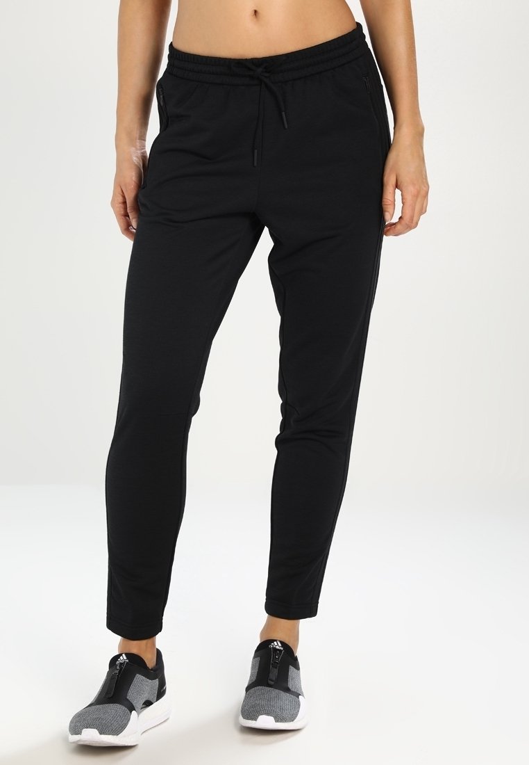 Žena v černých úzkých joggingových kalhotách s elastickým pasem a šedých atletických slip-on teniskách, stojící na bílé podlaze.