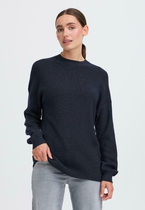 Pull en tricot bleu marine avec un col haut et des manches longues, légèrement oversize, arborant un motif texturé et des fentes latérales au niveau de l'ourlet.
