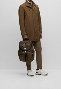 Mies, joka on pukeutunut ruskeaan takkiin ja housuihin, pitää kädessä ruskeaa reppua, jossa on useita taskuja, seisten harmaata taustaa vasten. Valkoiset lenkkarit näkyvät.