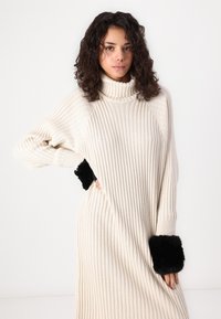 Cream ribbed sweater dress with a high neck, featuring black faux fur cuffs at the wrists. Relaxed fit with a textured fabric.