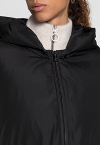 Schwarze Kapuzenjacke mit einer glatten, glänzenden Textur, Front-Reißverschluss mit einem runden Zipper und einem hellen, gerippten Kragen darunter.
