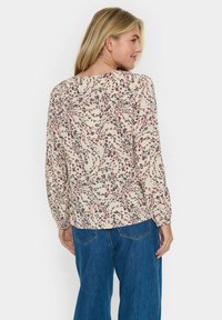 Blouse à motif floral en crème avec des accents rouge et bleu. Elle possède des manches longues, une coupe ample et un col rond. Finition avec des poignets élastiques.
