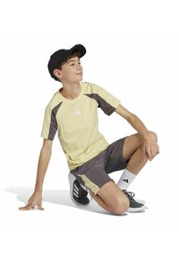 Gelbes Sport-T-Shirt mit dunkelgrauen Akzenten und Logo, kombiniert mit grauen Shorts, sitzende Pose, trägt eine schwarze Cap und Sneakers.