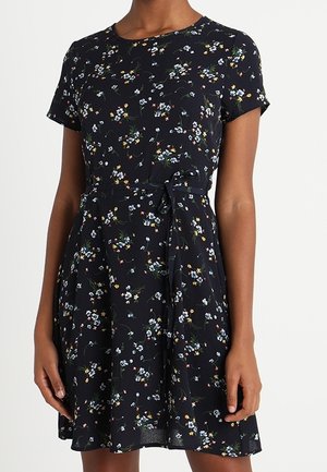 Mujer con un vestido negro de manga corta con un pequeño patrón floral multicolor y un lazo en la cintura, posando contra un fondo blanco.