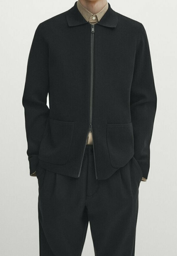 Personne portant une veste noire zippée avec deux poches avant sur une chemise marron clair à col et un pantalon noir, sur un fond clair uni.