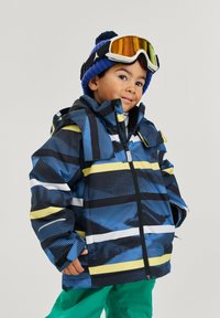Blau- und schwarz gemusterte Jacke mit gelben und weißen Streifen, einem Reißverschluss und einer Kapuze. Kombiniert mit grünen Hosen und einer blauen Mütze.