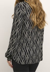 Femme avec de longs cheveux ondulés portant un blouse noire avec des motifs verticaux crème en forme de liane et un pantalon noir, vue de dos.