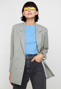 Tarkastettu bleiseri vihreänä, sinisenä ja valkoisena; kaksirivinen malli; yhdistetty siniseen t-paitaan ja korkeavyötäröisiin mustiin farkkuihin; pyöreät keltaiset aurinkolasit.