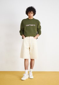 Felpa verde oliva con logo "carhartt." bianco, abbinata a pantaloni corti color crema. Indossata con sneakers bianche e calzini bianchi.