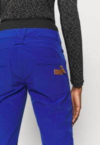 Niebieskie spodnie o gładkiej fakturze, wyposa�żone w dwie tylne kieszenie oraz małą brązową skórzaną metkę. Pas jest czarny i elastyczny.