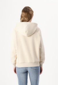 Donna con capelli castano medio che indossa un hoodie beige e jeans azzurri chiari, in piedi di fronte a uno sfondo bianco con le mani rilassate.