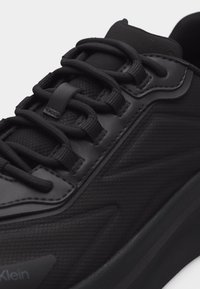 Κοντινό πλάνο μαύρου αθλητικού παπουτσιού που αναδεικνύει υφασμάτινη υφή, παχιά κορδόνια, επενδεδυμένη γλωττίδα και ραμμένα συνθετικά επικάλυψης στο πλάι.