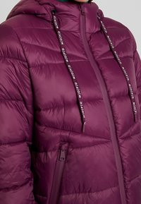 Fioletowa kurtka puchowa z kapturem, z poziomym przeszyciem, pełnym zamkiem błyskawicznym i dekoracyjnymi sznurkami. Gładki, błyszczący materiał.