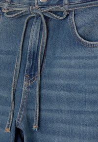 Gros plan sur un jean en denim bleu avec une ceinture en tissu nouée à la taille et des détails visibles de la poche avant et des boutons.