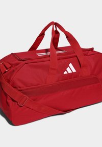 Bolso de viaje rojo con cierre de cremallera, asas reforzadas y correa de hombro ajustable. Presenta un logo y rayas blancas en la parte superior.