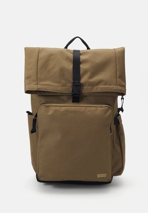 Sac à dos en tissu marron avec poche avant zippée, poches latérales, rabat supérieur sécurisé par une boucle noire et une poignée de transport sur le dessus.