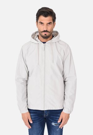 Uomo con capelli scuri e barba, indossa una giacca grigia chiara con cappuccio e cerniera e jeans blu strappati, in piedi contro uno sfondo bianco.