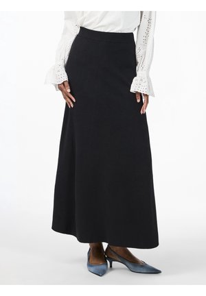 Donna con gonna lunga nera, camicetta bianca con occhielli e polsini svasati, e scarpe blu con tacco basso e punta affusolata su sfondo bianco.