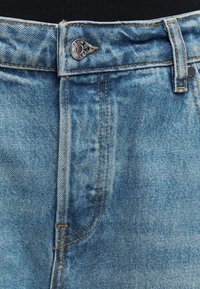 Κοντινή λήψη ανοιχτόχρωμων τζιν με μεταλλικό κουμπί με την επιγραφή «DENIM» και ορατές πορτοκαλί ραφές γύρω από τη μέση και το φερμουάρ.