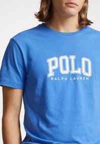 Blå bomulls t-shirt med en vit "POLO RALPH LAUREN" logga tryckt över bröstet, korta ärmar och rundad halsringning.