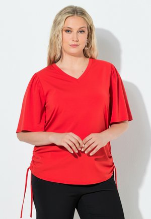 Blond kvinde iført rød V-hals top med rynkede sider og sorte bukser, stående mod en hvid baggrund med hænderne foldet.