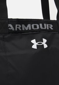 Bolso tote negro con tejido duradero, que presenta un logotipo blanco de Under Armour y una cinta negra de marca en la parte superior. Diseño simple sin patrones.