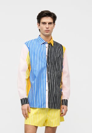 Jauns vīrietis valkā daudzkrāsainu ar vertikālām svītrām rotātu kreklu zilos, melnos, dzeltenos un rozā sadaļās, kā arī dzeltens svītrains šorti.