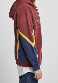 Sweat à capuche marron avec un cordon de serrage, présentant des accents diagonaux jaunes et bleu marine. Le tissu semble doux avec des poignets et un bas côtelés.