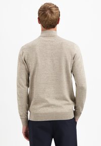 Mann, der von hinten steht und einen beigen langärmeligen Pullover sowie dunkle marineblaue Hosen trägt, mit den Händen in den Taschen vor einem weißen Hintergrund.