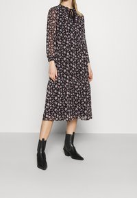 Robe midi noire à fleurs avec manches longues transparentes, ourlet volanté et col noué. Portée avec des bottines noires. Présente de petites fleurs roses.