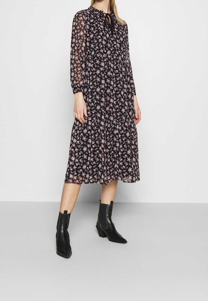 Robe midi noire à fleurs avec manches longues transparentes, ourlet volanté et col noué. Portée avec des bottines noires. Présente de petites fleurs roses.