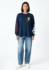 Femme portant un haut à manches longues bleu marine avec un éclair jaune, un texte rouge sur la manche gauche, un motif de flamme blanche sur la manche droite, un jean bleu clair et des baskets blanches.