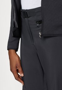 Pantalones técnicos negros con un diseño ajustado, que presentan una cinturilla visible con hebilla y un detalle de bolsillo con cremallera. Textura suave.