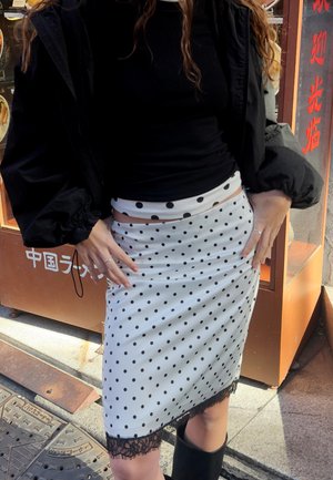 Donna che indossa una maglia nera a maniche lunghe e una gonna bianca a pois con bordatura in pizzo nero, in piedi vicino a una vetrina di un ristorante.