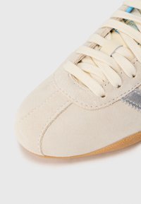 Lys beige semsket sneakers med hvite snørebånd, myk tekstur, avrundet tå og en kontrastgrå stripe langs siden. Gummisåle.