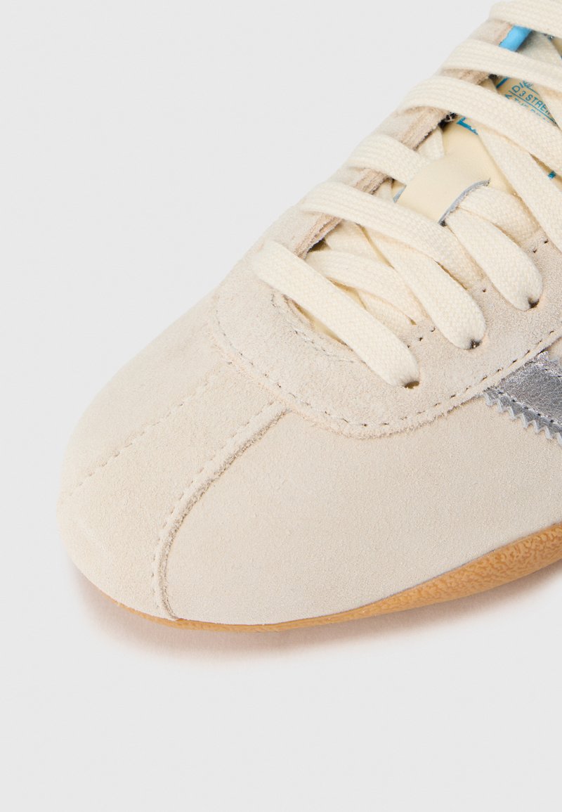 Sneaker in suede beige chiaro con lacci bianchi, texture morbida, punta arrotondata e una striscia grigia a contrasto lungo il lato. Suola in gomma.