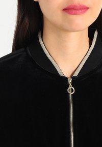 Černá sametová bomber bunda se zlatým akcentovaným zipem a kovovým límcem. Texturovaná tkanina a elegantní design zvýrazňují oděv.